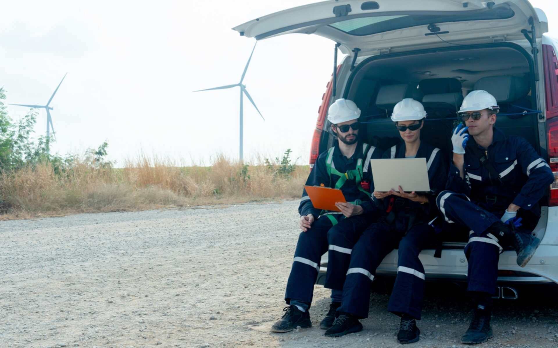 Three energy engineers review energy management contracts on a laptop near wind turbines.